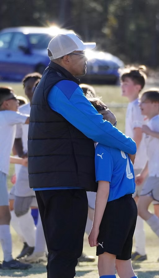 Coach G on the sideline with his arm around a young player in a blue jersey