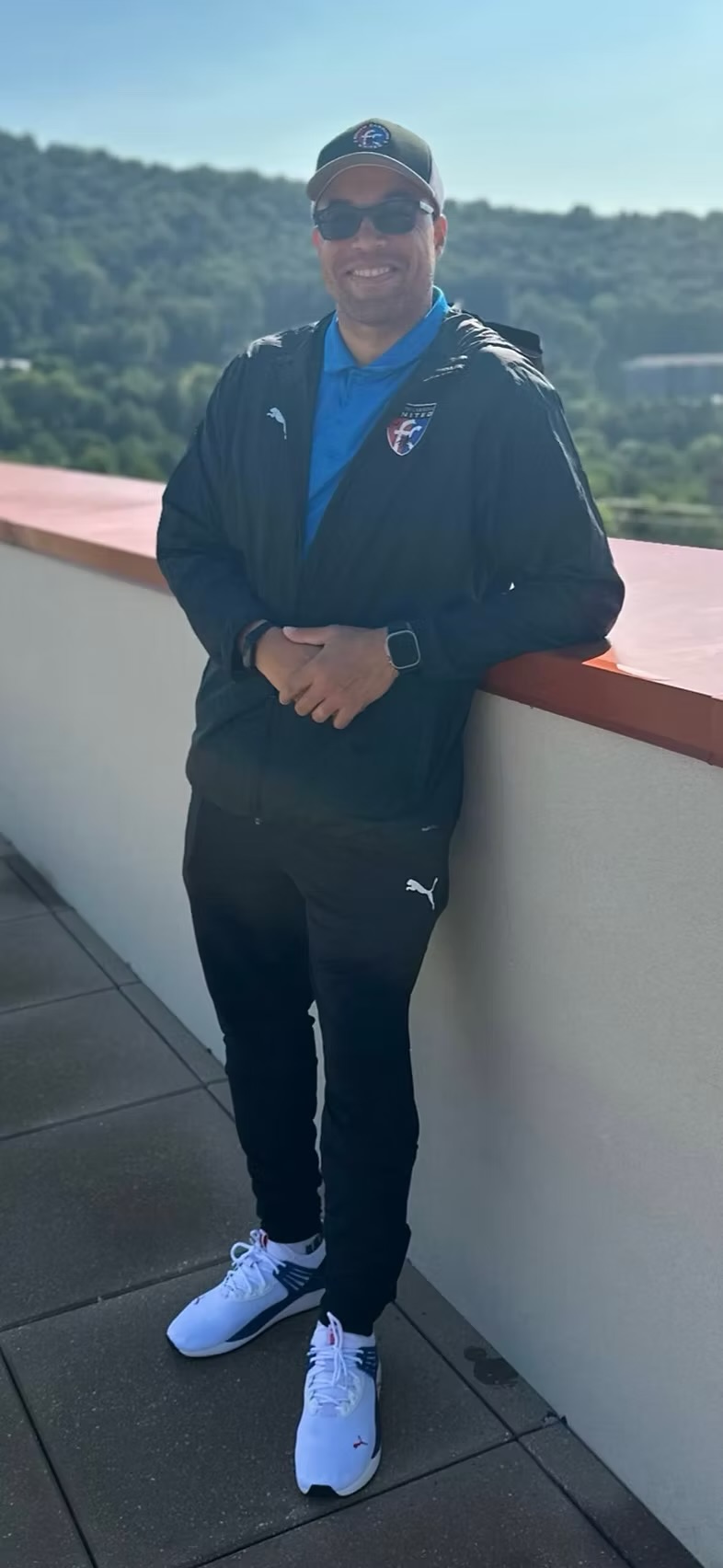 Coach G smiling on a rooftop terrace in a club jacket and cap, with wooded hills behind him